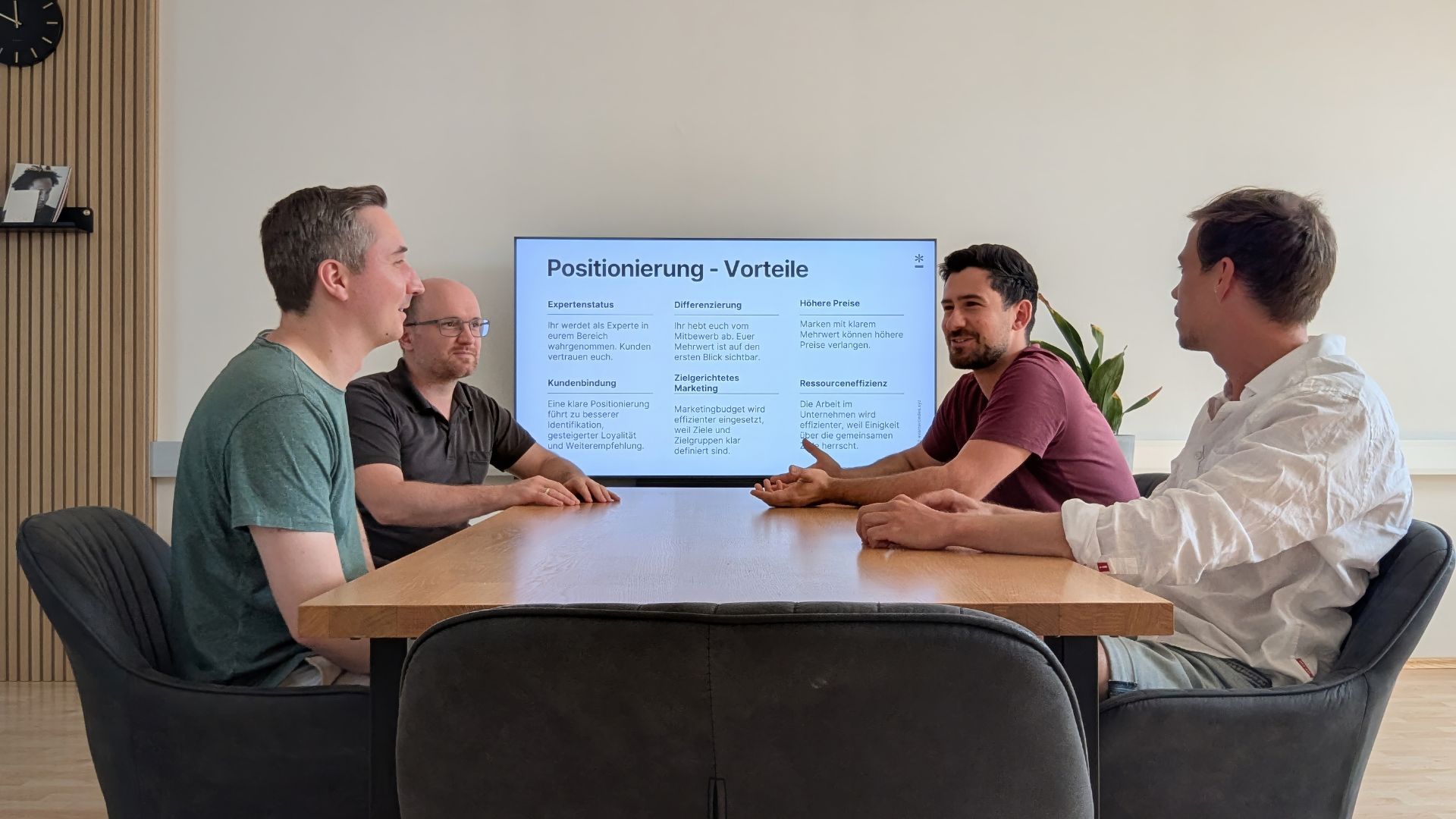 Vier männliche Gründer sitzen an einem Meeting-Tisch und unterhalten sich. Im Hintergrund ist ein Bildschirm zu sehen auf dem eine Präsentationsfolio zum Thema Positionierung angezeigt wird.