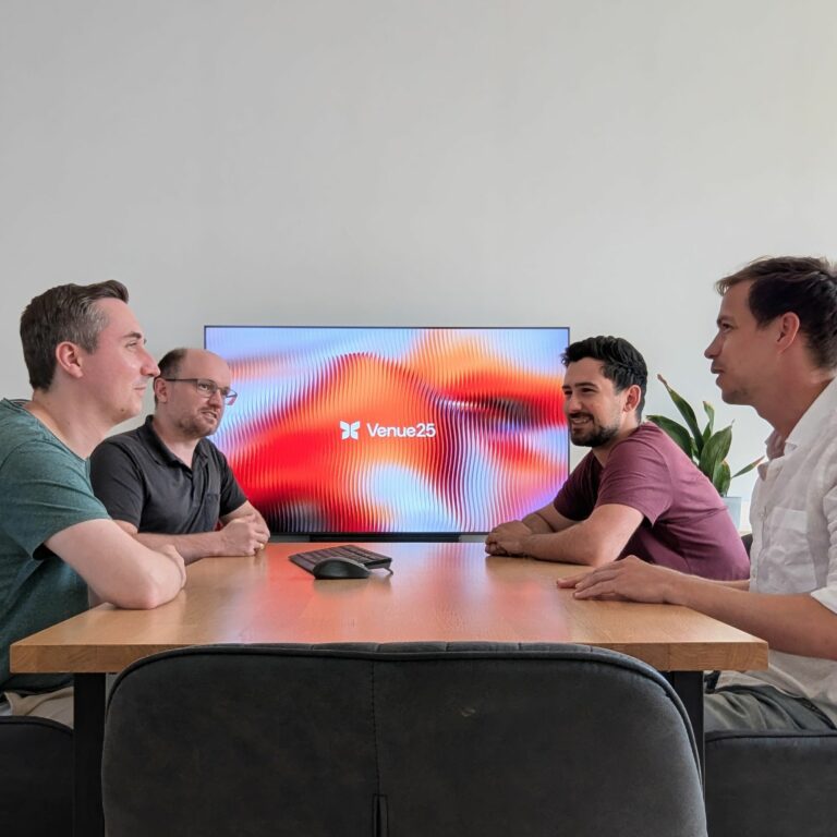 Vier männliche Gründer sitzen an einem Meeting-Tisch. Im Hintergrund ein Bildschirm, dass das Firmenlogo von Venue25 zeigt.